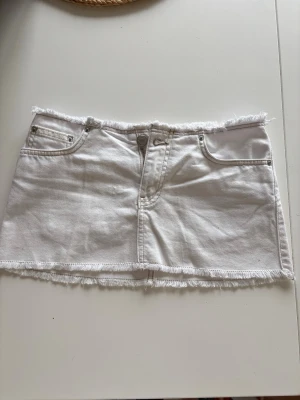 Vit jeanskjol med fransad kant midje mått 36 - Säljer en vit kort jeanskjol med fransade kanter och klassiska fem fickor. Kjolen har dragkedja och knapp framtill samt råa sömmar för en avslappnad look. Perfekt till sommaren och lätt att matcha med olika toppar.