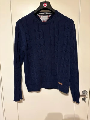 Mörkblå kabelstickad tröja Bondelid - Mörkblå kabelstickad tröja från Bondelid med rund halsringning och ribbade muddar. Tröjan har ett klassiskt flätmönster och en liten läderdetalj vid nederkanten. Perfekt för dig som gillar stilrena och tidlösa plagg.