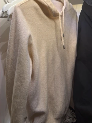 Beige cashmere hoodie - Säljer en stilren beige hoodie med huva och dragsnören. Hoodien har lång ärm och ribbade muddar vid ärmslut och nederkant. Tillverkad i cashmere!