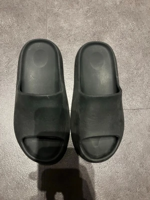 Svarta chunky slides i gummi - Säljer ett par svarta slides med chunky sula och öppen tå. Tillverkade i mjukt gummi för maximal komfort. Perfekta för chill dagar eller att glida runt hemma. Minimalistisk design utan synligt märke och räfflad undersida för bra grepp.