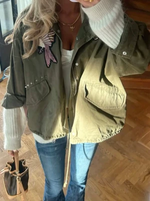 Olivgrön jacka med tryck - Snygg olivgrön vindjacka med broderade detaljer som fågel och rosor. Jackan har två stora fickor framtill, dragsko i nederkant och tryckknappar. Oversized passform och coola nitar på ena fickan ger en trendig look. Perfekt för dig som vill sticka ut.