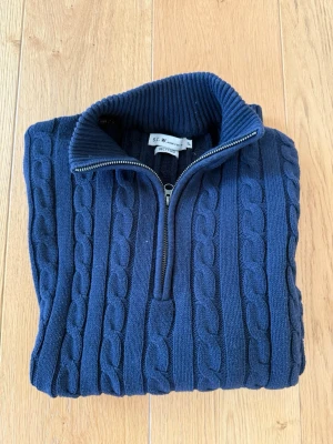 Mörkblå kabelstickad tröja med halvzip - Mörkblå kabelstickad tröja från S.C. W. Recycled med halvzip och hög krage. Tröjan har långa ärmar, ribbade muddar och klassiskt kabelmönster. Perfekt för dig som gillar en stilren och tidlös look.