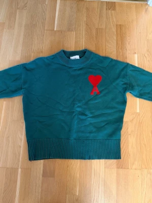 Grön stickad tröja från Ami Paris - Mörkgrön stickad tröja från Ami Paris med röd hjärtlogga och bokstaven A på bröstet. Tröjan har rund halsringning, ribbade muddar och nederkant. Perfekt för dig som gillar stilrena detaljer och vill ha något som sticker ut.