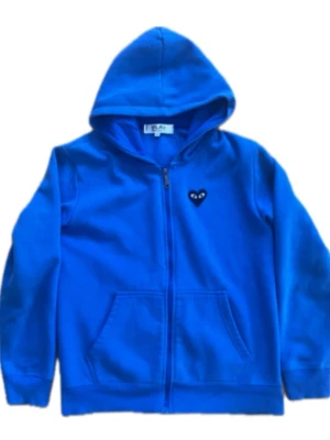 Blå hoodie från Comme des Garçons - Blå hoodie med dragkedja från Comme des Garçons Play. Ikoniskt hjärta med ögon på bröstet och stort hjärta med ögon på ryggen. Tillverkad i mjuk bomull, har huva och kängurufickor framtill. OBS. tveka inte på att höra av er vid frågor!