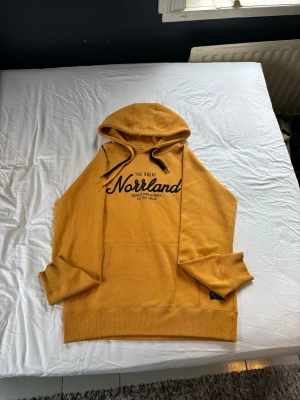 Gul hoodie The Great Norrland - Gul/Orange hoodie från The Great Norrland med svart tryck på bröstet och stor huva med dubbla snören. Klassisk känguruficka framtill och ribbade muddar vid ärmar och nederkant. Jag är ca 180cm och väger 82kg sitter lite tajt på mig men passar defenitvt någon som är lite mindre