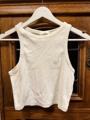 Vit ribbad croppad linnetopp HM - Säljer en enkel och stilren vit croppad linnetopp i ribbad bomull. Toppen är ärmlös med breda axelband och har en klassisk rund halsringning. Perfekt att matcha med jeans eller kjol för en clean look.