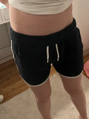 Svarta shorts med vit kant och snörning - Säljer ett par svarta shorts med vit kant och vit snörning i midjan. Shortsen har elastisk midja och är perfekta för sommarens alla aktiviteter. Sportig och avslappnad stil. Från cubus 