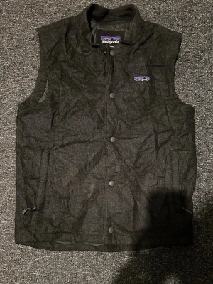 Patagonia väst - Snygg mörkgrå dunväst från Patagonia med hög krage och tryckknappar framtill. Västen har två sidofickor med dragkedja och klassisk Patagonia-logga på bröstet. Perfekt lager på lager och riktigt skön att bära under kyliga dagar.