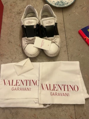 Valentino Garavani vita sneakers - Snygga vita sneakers från Valentino Garavani med svart band över vristen och klassisk logga på plösen. Skorna har en chunky sula och är tillverkade i läder. Medföljer originalpåse och tag. Perfekta för dig som vill ha en lyxig och trendig look.