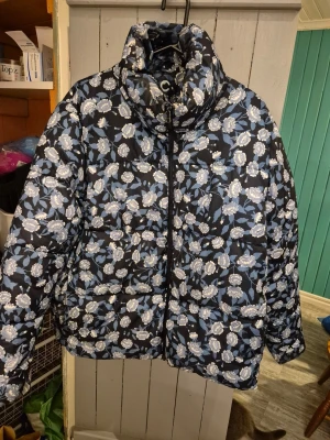 Pufferjacka med hög krage - Snygg pufferjacka med mönster i blått och vitt. Jackan har hög krage, dragkedja framtill och två fickor. Perfekt för kalla vinterdagar och ger en cool, trendig look. Materialet är glansigt och quiltat för extra värme.