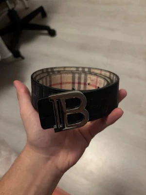Burberry vändbart bälte med B-spänne - Snyggt vändbart bälte från Burberry med klassiskt rutigt mönster på ena sidan och svart på andra. Stort metallspänne med B-logga framtill. Perfekt accessoar för att lyfta din outfit. Tillverkat i skinn.
