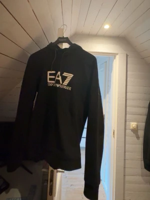 Svart hoodie från Emporio Armani EA7 - Snygg svart hoodie från Emporio Armani EA7 med stor vit logga på bröstet. Klassisk huva med dragsko och magficka framtill. Tillverkad i mjukt material som känns skönt mot huden. Perfekt för en avslappnad och trendig look.