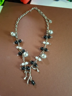Silverfärgat halsband med svarta och vita pärlor - Unikt halsband med silverfärgad kedja dekorerad med stora och små svarta och vita pärlor. Pärlorna sitter i kluster längs kedjan och ger ett coolt och trendigt uttryck. Halsbandet har även en dekorativ detalj längst ner som ger extra edge.