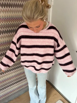 Rosa och brun randig stickad tröja - Mysig stickad tröja i ljusrosa med breda svarta ränder. Tröjan har en avslappnad passform, rund halsringning och långa, lite pösiga ärmar. Perfekt för dig som gillar en chill och trendig look. Materialet ser ut att vara mjukt och fluffigt. Xl men sitter som s/m