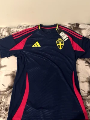 Sverige fotbollströja Adidas blå M - Säljer en officiell blå Sverige fotbollströja från Adidas med gula och rosa detaljer. Tröjan har korta ärmar, v-ringning och Sveriges landslagsemblem på bröstet. Tillverkad i polyester och perfekt för fotboll eller att bära till vardags.