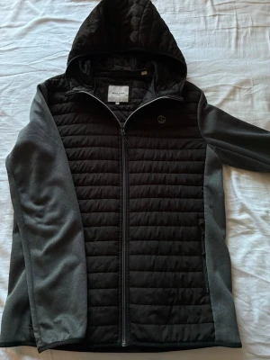 Svart vindjacka från Jack & Jones - Snygg svart vindjacka från Jack & Jones med quiltad front och huva. Ärmar och sidor i mörkgrått mjukt material, hel dragkedja och två sidofickor med dragkedja. Perfekt för blåsiga dagar och enkel att matcha med olika outfits.