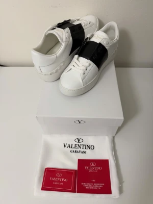 Valentino Garavani vita sneakers - Snygga vita sneakers från Valentino Garavani med en bred svart läderdetalj över vristen. Skorna har klassisk låg profil, rund tå och vita snören. Tillverkade i läder med subtila nitar på hälen för extra edge. Kommer med originalkartong och dustbag.