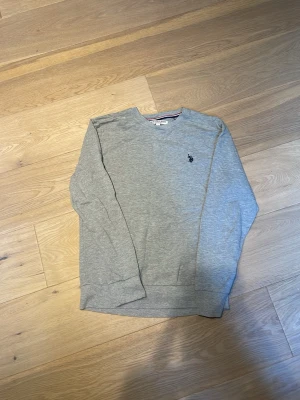 Grå sweatshirt från U.S. Polo Assn. - En stilren grå sweatshirt från U.S. Polo Assn. med rund halsringning och diskret logga broderad på bröstet. Tröjan har långa ärmar och ribbade muddar vid ärmslut och nederkant. Perfekt för en avslappnad look.