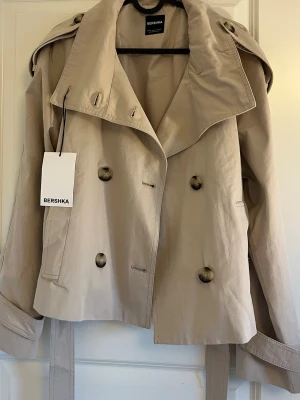 Kort beige trenchcoat från Bershka - Stilren kort trenchcoat från Bershka i färgen  beige. Helt oanvänd med prislapp kvar