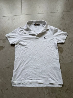 Vit pikétröja från Ralph Lauren - Klassisk vit pikétröja från Ralph Lauren med blå broderad logga på bröstet. Tröjan har korta ärmar, krage och knappar vid halsen. Tillverkad i bomull för en skön och luftig känsla. Perfekt till jeans eller shorts.