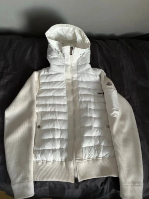 Vit Moncler Cardigan - Säljer en snygg vit pufferjacka från Moncler med quiltad framsida och huva. Jackan har stickade ärmar och sidor, dragkedja framtill och Moncler-logga på ärmen. Perfekt för dig som vill ha en clean och stilren look under kyliga dagar. Inga defekter förutom en liten ”fläck” som knappt syns(Bild 4)