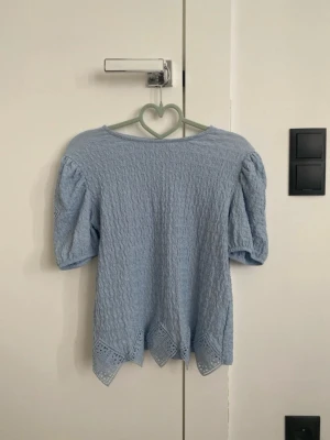 Ljusblå blus med spetsdetaljer - Supersöt ljusblå blus med puffiga korta ärmar och vågig spetskant längst ner. Materialet är strukturerat och ger en unik look. Perfekt för dig som vill ha något gulligt och stilrent i garderoben.