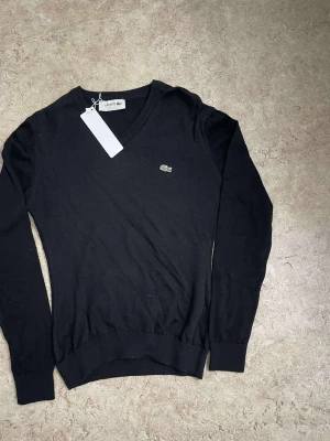 Svart V-ringad tröja från Lacoste - Klassisk svart stickad tröja från Lacoste med V-ringning och den ikoniska gröna krokodilloggan på bröstet. Tröjan har långa ärmar och ribbade muddar vid ärmslut och nederkant. Perfekt för en stilren och enkel look.
