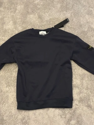 Mörkblå sweatshirt från Stone Island - Snygg mörkblå sweatshirt från Stone Island med klassisk rund hals och ribbade muddar. Tröjan har den ikoniska logotypen på ärmen och är tillverkad i mjuk bomull för skön komfort. Perfekt för en clean och stilren look.