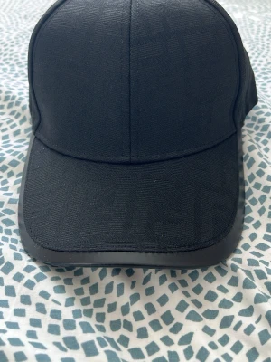 Svart keps från Fendi - Snygg svart keps från Fendi med klassisk böjd skärm och diskret struktur i tyget. Insidan har Fendi-loggor på banden och justerbar rem bak med gulddetalj. Perfekt accessoar för en clean och stilren look.