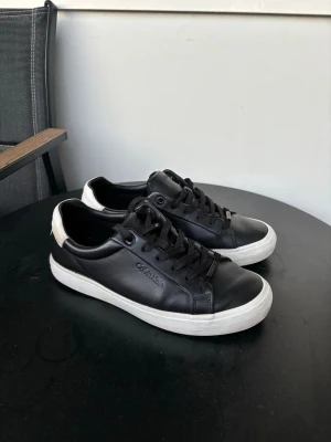 Svarta Calvin Klein sneakers i läder - Snygga svarta sneakers från Calvin Klein i slätt läder med vita sulor och svart snörning. Diskret logga på sidan och kontrastdetalj i vitt på hälen. Perfekta för en clean och stilren look.