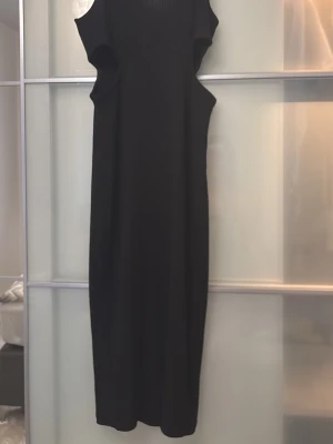 Svart långklänning med cut out - Stilren svart långklänning med cut out-detaljer vid midjan och axelbandslös design. Klänningen har en smickrande, rak passform och är perfekt för dig som vill ha en elegant look. Materialet känns mjukt och följsamt.