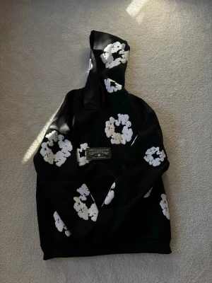 Svart hoodie med vita blommor Denim Tears - Säljer en svart hoodie från Denim Tears med stora vita blommor tryckta över hela plagget. På bröstet sitter en patch med texten 'African Diaspora Goods Denim Tears'. Hoodien har huva och är gjord i mjukt bomullsmaterial.