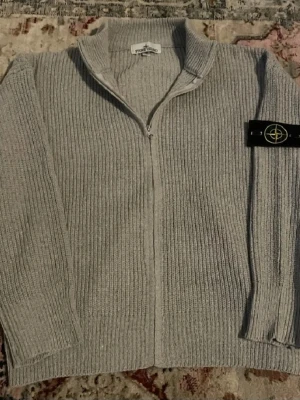 Grå ziptröja från Stone Island - Säljer en grå ribbstickad ziptröja från Stone Island med klassisk logotyp på ärmen. Tröjan har hög krage och dragkedja framtill. Perfekt för lager-på-lager och streetwear-stil.