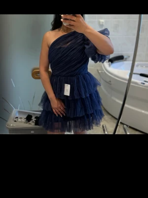 Marinblå oneshoulder volangklänning ByMalina - Säljer en marinblå kortklänning med oneshoulder och flera lager volanger. Klänningen har en asymmetrisk axel och är tillverkad i ett tunt, lätt material som ger en fluffig look. Perfekt för dig som vill sticka ut med en unik och trendig stil.