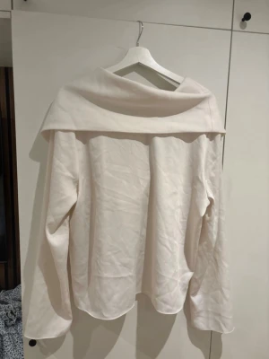 Vit omlottopp med öppen rygg och rosett - Trendig vit topp med bred omlottkrage och öppen rygg, dekorerad med en söt rosett baktill. Långärmad och i mjukt tyg som ger en avslappnad men chic look. Perfekt för dig som vill sticka ut med en unik detalj på ryggen.