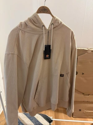 Beige hoodie från Dickies i manchester - Säljer en beige hoodie från Dickies i mjuk manchester med huva och dragsko. Hoodien har en stor magficka och diskret Dickies-logga på fickan. Oversized passform och ribbade muddar vid ärmslut och nederkant. Perfekt för en chill och avslappnad stil. (OG pris var 650kr)