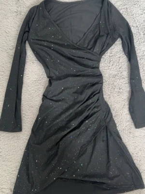 Svart glittrig omlottklänning - Snygg svart omlottklänning med glitterdetaljer och v-ringning. Klänningen är från Shein eller temu. Använts en gång på nyår, annars är den alldrig använd. 