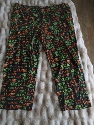 Xbox Live pyjamasbyxor - Säljer ett par svarta pyjamasbyxor med färgglatt Xbox Live-tryck i grönt och orange över hela tyget. Byxorna har resår i midjan och är perfekta för gamingkvällar eller chill hemma. Materialet känns mjukt och bekvämt mot huden.
