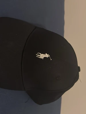 Svart keps från Polo Ralph Lauren - Snygg svart keps från Polo Ralph Lauren med den klassiska broderade loggan i vitt framtill. Kepsen har böjd skärm och justerbar rem bak. Perfekt accessoar för en stilren och sportig look.