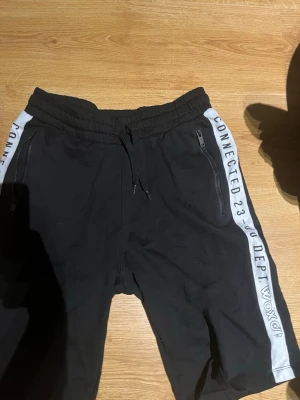 Svarta shorts från Woxo med tryck - Svarta shorts från Woxo med vita sidopaneler och texttryck längs sidorna. Shortsen har snörning i midjan och dragkedjeförsedda fickor framtill. Perfekta för en avslappnad och sportig stil.