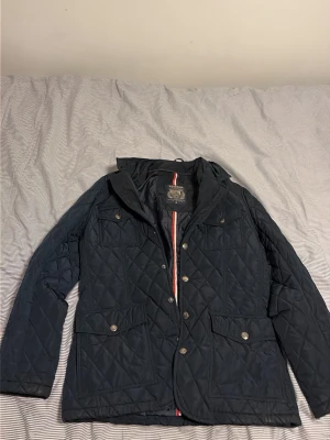 Mörkblå quiltad jacka från Barbour - Snygg mörkblå quiltad jacka från Barbour med klassisk krage, tryckknappar och dragkedja. Jackan har fyra fickor framtill och diskreta detaljer i rött och vitt längs dragkedjan. Perfekt för kyliga dagar eller sköna sommarkvällar