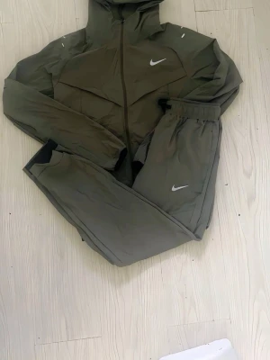 Nike olivgrön träningsset herr - Säljer ett olivgrönt träningsset från Nike med matchande jacka och byxor. Setet har dragkedja framtill, huva och elastisk midja på byxorna. Båda plaggen har vita Nike-loggor och är tillverkade i ett lätt, sportigt material. Perfekt för träning eller chill.