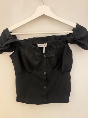 Svart offshoulder topp från Kooliok - Trendig svart offshoulder topp från Kooliok med knappar framtill och rynkade detaljer vid bysten. Toppen har korta puffärmar och en croppad passform som ger en snygg siluett. Perfekt att styla med höga jeans eller kjol.