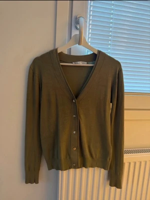 Olivgrön kofta med v-ringning - Snygg olivgrön kofta med v-ringning och knappar framtill. Långärmad modell i tunt, mjukt material som passar perfekt till lager på lager. Enkel och stilren design utan mönster.