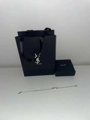 Y.S.L - Snyggt och stilrent armband silverfärgad metall. Armbandet har en tunn kedja och en liten Y.S.L logga som detalj. Perfekt accessoar för att lyfta din outfit med en lyxig touch. 