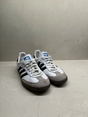 Adidas Samba vita och svarta sneakers - Klassiska Adidas Samba sneakers i vitt med svarta ränder och grå mockadetaljer på tån. Skorna har en låg profil, vit ovandel i läder och gummisula. Ikonisk blå Adidas-logga på plösen och svarta detaljer på hälen. Perfekta för en sportig och stilren look.