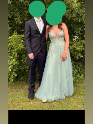 Ljusgrön långklänning med spetsdetaljer - Säljer en elegant ljusgrön långklänning med v-ringning och tunna axelband. Klänningen har vackra spetsapplikationer och ett skirt tylltyg som ger en romantisk känsla. Perfekt för bal eller student. Kjolen har en slits framtill för extra stil. (Finns slips och näsduk i samma färg om man vill ha) 