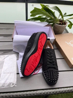 Svarta Louboutin sneakers med nitar - Säljer ett par svarta sneakers från Christian Louboutin med klassisk röd sula och coola svarta nitar på tån. Skorna har snörning och är tillverkade i mocka för en lyxig känsla. Perfekta för dig som vill sticka ut med en edgy och exklusiv stil.