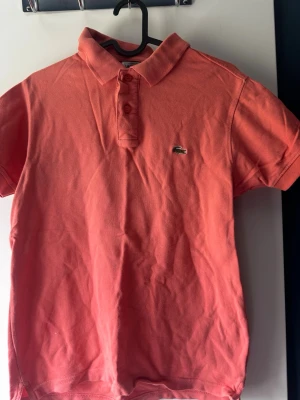 Korallröd piké från Lacoste - Säljer en klassisk korallröd pikétröja från Lacoste med korta ärmar och den ikoniska krokodilloggan på bröstet. Tröjan har krage och knappar framtill, tillverkad i mjuk bomull som andas. Perfekt för en avslappnad och stilren look.