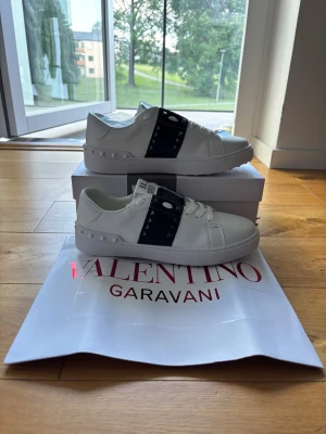 Valentino Garavani vita sneakers - Snygga vita sneakers från Valentino Garavani med svart band och nitar på sidorna. Skorna är låga och har en chunky sula samt coola detaljer i läder. Perfekta för dig som vill ha en stilren men edgy look.